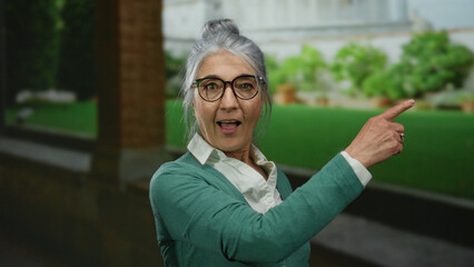 Senior woman with grey hair wearing glasses gives a thumbs up gesture while standing in an outdoor...