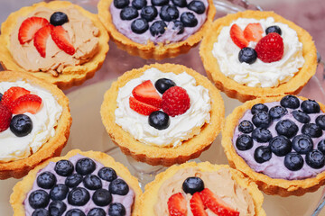 Mini tartlets with cream and fresh berries. Colorful fruit desserts with blueberries, strawberries and raspberries. Sweet bakery pastry on glass plate. Delicious homemade treats for party.