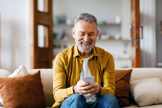 Senior man using smartphone texting, networking in social media or scrolling social media, sitting on sofa at home. Gadgets and retirement lifestyle