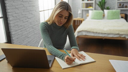 Woman writing with a pen in a notebook on a desk beside a laptop in a cozy bedroom, showcasing focused home study or work environment with green cushions on the bed.