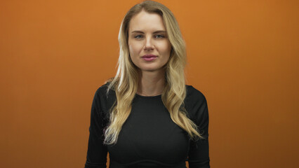 Woman with blonde hair in black shirt standing against a vibrant orange background, looking...