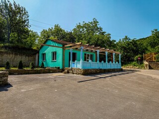 A small blue house with a white roof sits on a gravel driveway. The house is surrounded by trees and has a patio area with a pergola