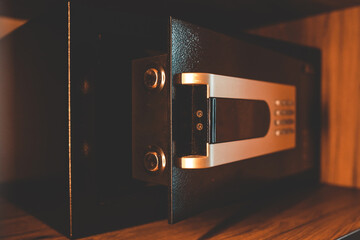 Close-up of modern digital safe with open door inside wooden hotel closet. Secure storage box for valuables with keypad and metal handle in warm lighting