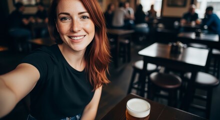 A smiling young woman with red hair takes a selfie in a cozy bar, holding a glass of beer on the table in front of her.