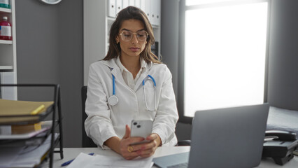 Woman doctor working in hospital room, wearing glasses and lab coat, using phone with laptop on...