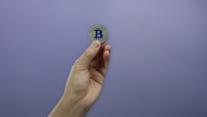 Caucasian man hand holding bitcoin coin against purple wall background, highlighting cryptocurrency...