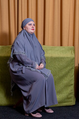 A Muslim woman in a modest gray hijab sits against an orange curtain