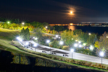 A peaceful city park at night with illuminated benches, soft streetlights, and moonlight reflecting on the river.