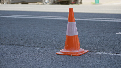 Traffic cone standing on sunlit urban street surrounded by moving vehicles and people nearby.