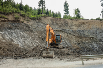 Excavator working at construction site near road. Heavy machinery digging soil on rocky slope with...
