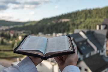 Person holding open Bible on balcony with mountain view and city buildings in background. Concept...