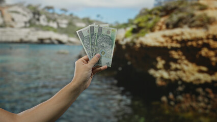 Hand holding polish zloty banknotes with a scenic seaside beach and water in the background under a clear sky, capturing a sunny outdoor financial moment.