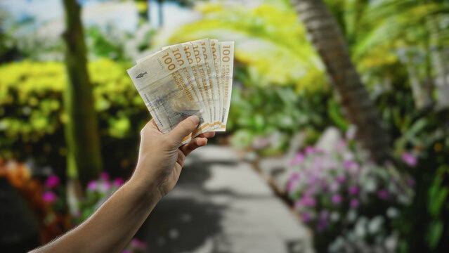 Man holding danish krone banknotes in hand outdoors amidst a lush garden setting, showcasing a vibrant park environment with blurred greenery in the background.