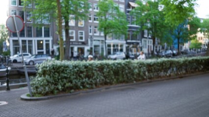 Urban scene in amsterdam with blurred buildings and people cycling, showcasing greenery, parked cars, and historic architecture under a bright sky.