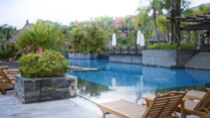 Blurred tropical resort scene with defocused wooden loungers by a luxury pool surrounded by lush green plants in an outdoor paradise setting.