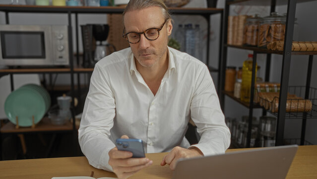 Mature caucasian man with glasses using smartphone and laptop at a wooden desk in a modern office with microwave and shelves in the background.