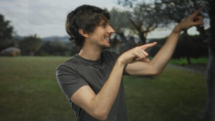 Young smiling man wearing gray tshirt finger pointing to tree in green forest; curiosity...