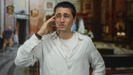 Obraz premium Young hispanic man in church expressing frustration indoors wearing white shirt with contemplative expression under soft lighting showing diverse emotions in a spiritual setting.