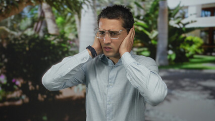 Young hispanic man wearing glasses covers ears with palms in park amid lush trees; annoyance frustration irritation distress.
