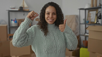 Young woman smiling holding keys in living room surrounded by boxes in her new home showing thumbs...
