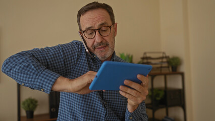 Mature man in office using tablet while talking on smartphone, showcasing multitasking in a...