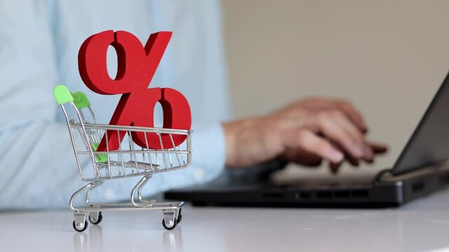 Red sign of percent in small shopping trolley on the table. Man using laptop on the background. Sales, Black Friday, online shopping concept.