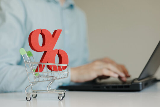 Red sign of percent in small shopping trolley on the table. Man using laptop on the background. Sales, Black Friday, online shopping concept.