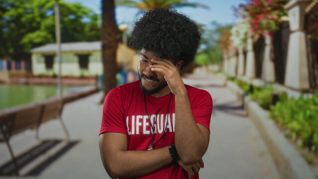 Man lifeguard outdoors in green park showing thoughtful expression with bench, trees in background wearing red shirt and watch during sunny day with blurred landscape.