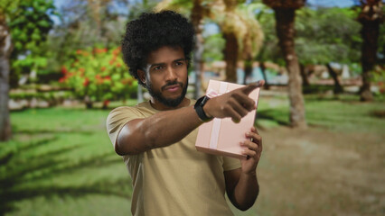 Man in park holding a pink gift box with ribbon in a green outdoor setting exhibiting a thoughtful...