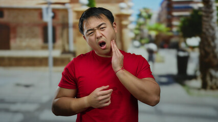Young man in red shirt suffers toothache on city street with blurred urban background, expressing...