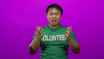 Asian man wearing green volunteer shirt looking emotional against vibrant pink isolated background,...