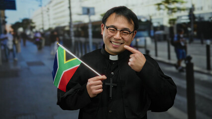 Priest holding south african flag on urban street with cheerful expression, wearing glasses and black clerical attire.