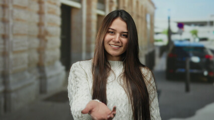 Woman standing outdoors on street smiling in casual white sweater with long hair, offering hand...