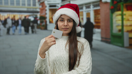 Woman wearing santa hat holds card on busy street showing holiday season shopping in urban setting.