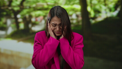 Woman in pink jacket outdoors in park setting with hands on face appearing thoughtful under lush...