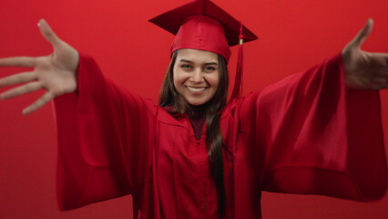 Woman smiling in graduation gown and cap on red background, extending arms in celebration and joy.