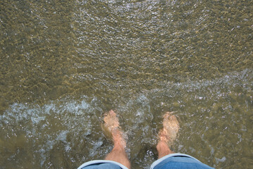 A first-person view shows bare feet standing in clear, shallow water with small waves gently...