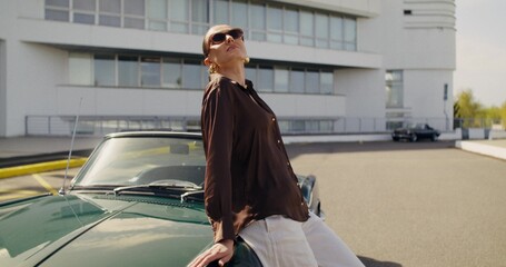 A stylish woman poses standing next to a retro convertible. Photo shoot on a sunny summer day