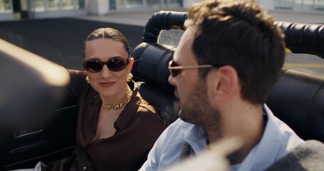 A stylish young man and woman are smiling and talking. They're sitting in a retro convertible on a...