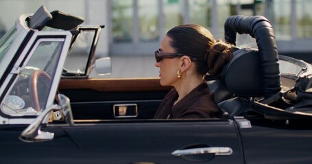 A stylish woman in sunglasses turns and looks at the camera while sitting in the driver's seat of a...