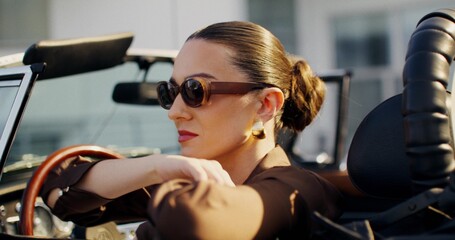 A stylish woman in sunglasses turns and looks out the window of a convertible. She's sitting in the...
