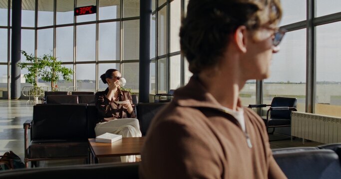 Stylish young woman in the airport lounge. She drinks coffee sitting in a chair next to other passengers.