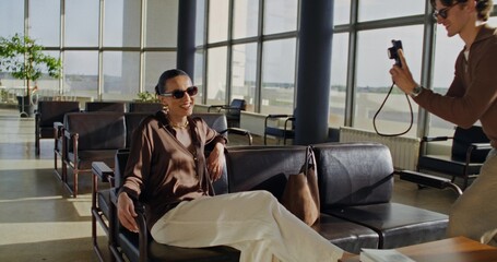 A man takes pictures of a woman on camera in the airport lounge. A woman poses sitting in an...