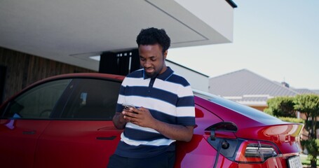 A man uses a mobile phone while standing near an electric car. He disconnects the charging cable...