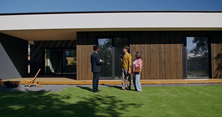 The realtor shows the house to the buyers. They are standing in the backyard and talking on a summer day.