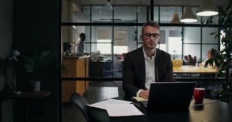 A businessman sits at a desk in front of an open laptop in the office. He smiles at the camera.