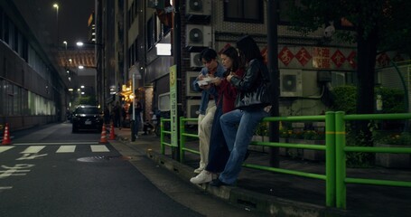 Two young women and a man are eating standing on a city street at night. They're talking and they look happy.
