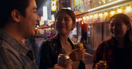 Funny young people spend time together. They drink beer and talk while standing on a city street at night.