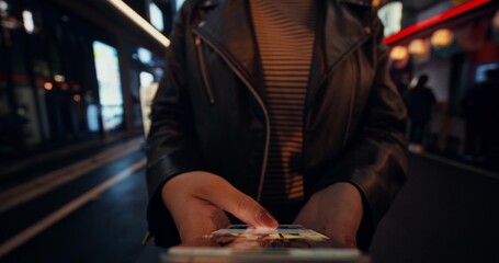 A woman uses a mobile phone while standing on a city street at night. Close-up of her hands, unrecognizable person