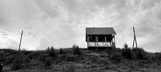 Black and white photo of a solitary wooden cabin on a hill against a cloudy sky. The scene conveys solitude and tranquility, with sparse vegetation.